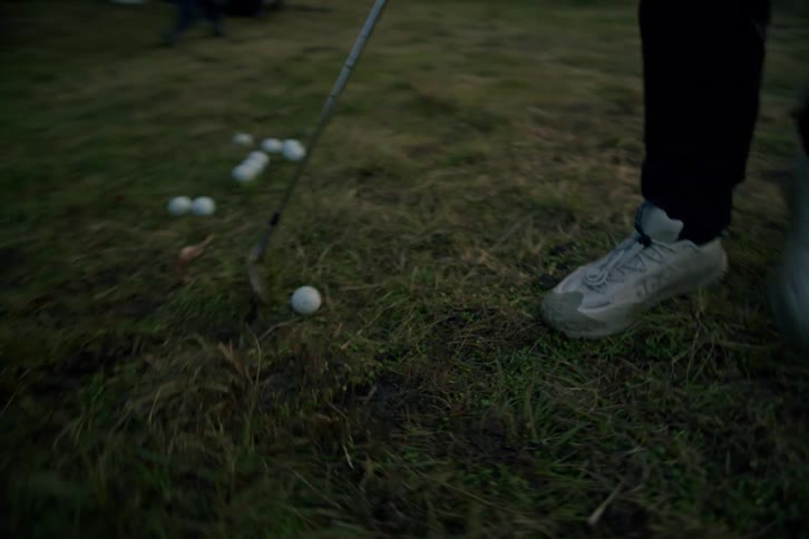A man walks across a grassy field at dusk, dragging his golf club towards a cluster of practice balls in slow motion