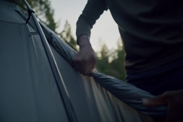 A man's hands carefully assemble a tent in the forest as the sun sets, casting a warm glow