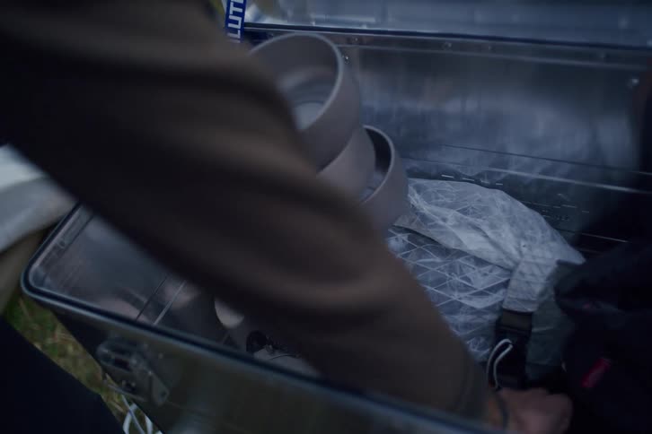 A person with tattooed arms carefully unpacks a lightweight titanium cook set from a silver aluminum storage box outdoors