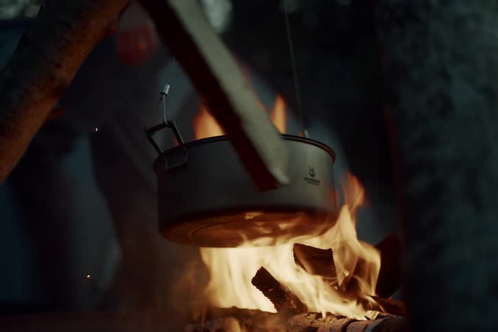 A metal pot hangs over a crackling campfire in slow motion with a tent in the background and a stick stirring the embers