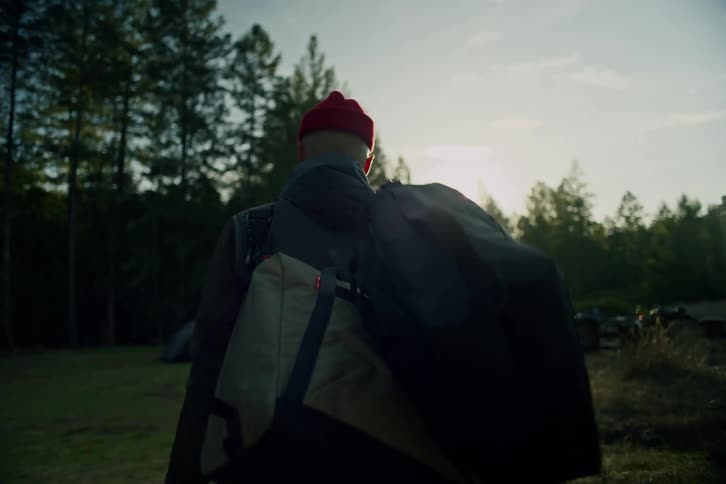 A man in a red beanie carries his camping gear through a sunlit field towards a forest at golden hour