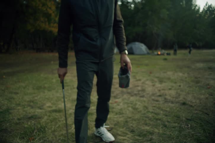 A man in outdoor gear practices his golf swing in slow motion at a rustic campsite in the woods