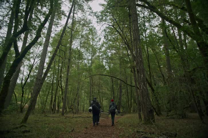 Two men carry fishing gear and a metal case down a trail deep into a lush green forest