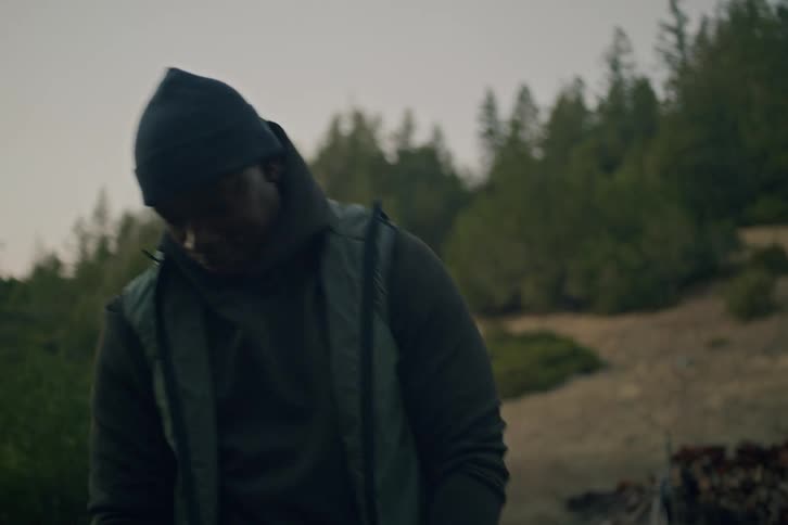 A man in a beanie practices his golf swing in slow motion in a rustic, natural field at dusk
