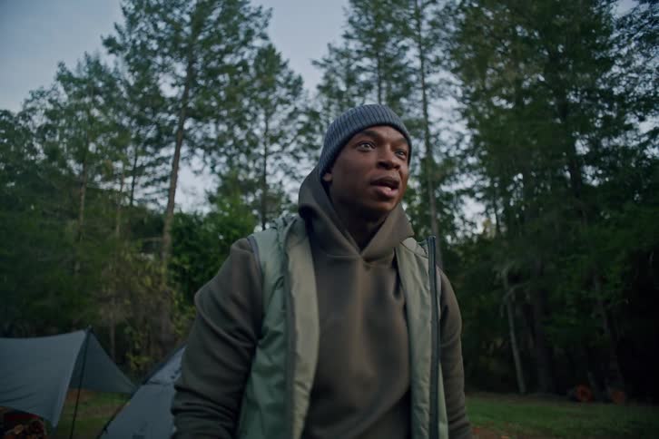 A man takes an impressive slow-motion golf swing at a campsite surrounded by trees as his friend watches on
