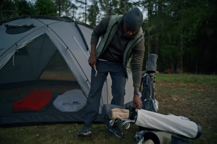 A happy man takes a golf club out of his bag while camping in the woods and walks towards a campfire