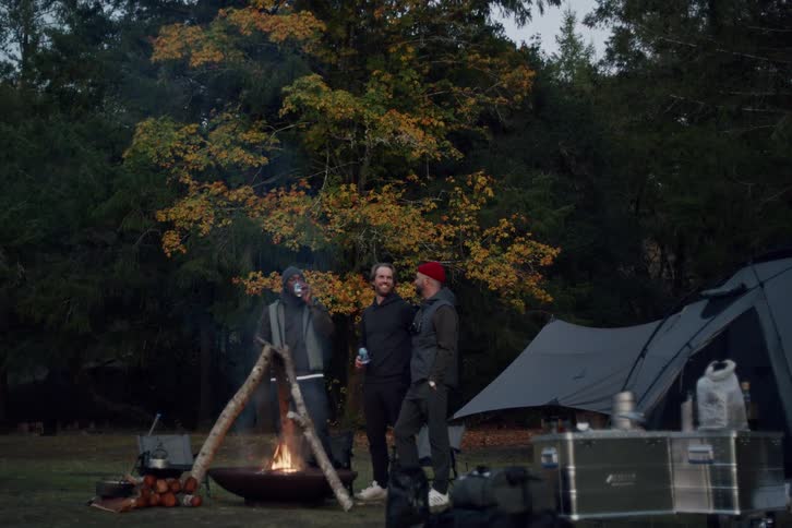 Three diverse male friends share a laugh while gathered around a warm campfire at their wooded campsite in the evening
