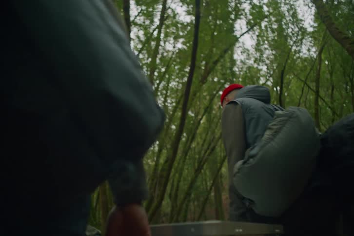 Two friends carry a large metal case together as they hike through a dense forest with their group