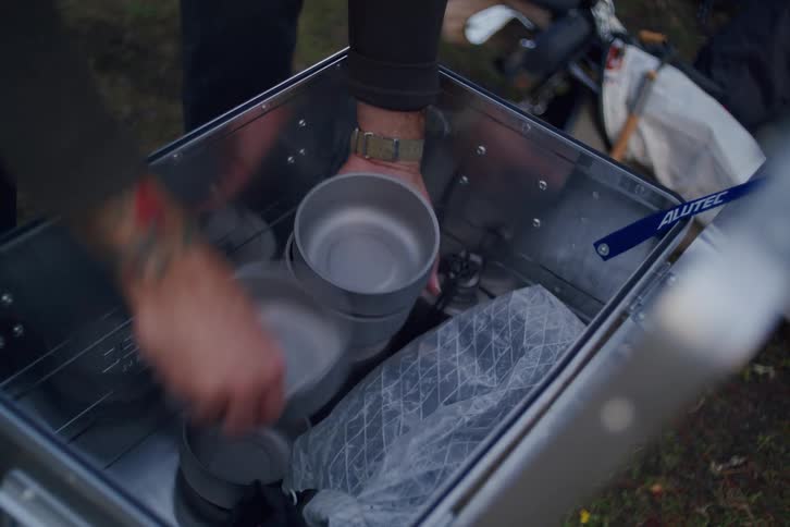 A tattooed adventurer unpacks a lightweight titanium cooking set from a metal storage case at a campsite