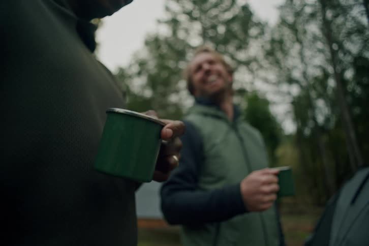 A close-up of a man's hand holding a green camping mug while his friend smiles in the background at their campsite in the woods