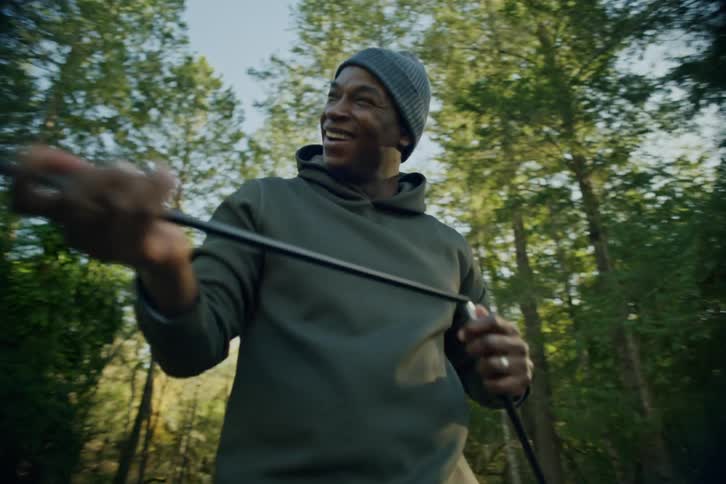 A joyful Black man wearing a beanie and hoodie smiles while assembling a tent pole in a sunny forest campground
