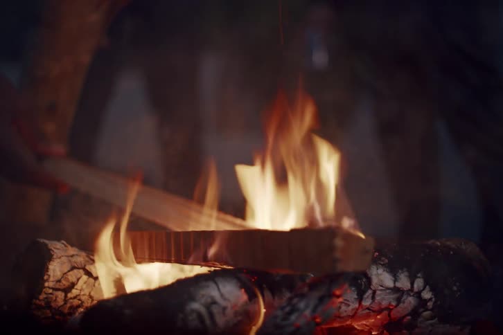 A person stokes a vibrant campfire with a piece of wood, causing embers and sparks to fly into the dark night in slow motion