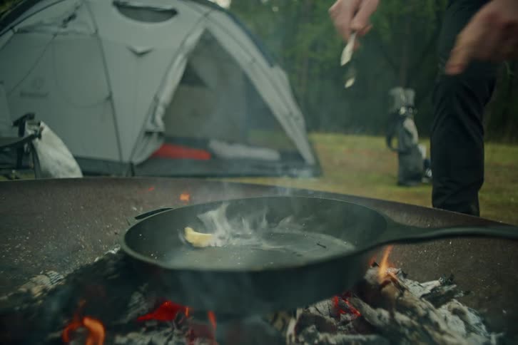 A pat of butter is dropped into a hot cast iron skillet over a campfire, melting and smoking instantly with a tent in the background
