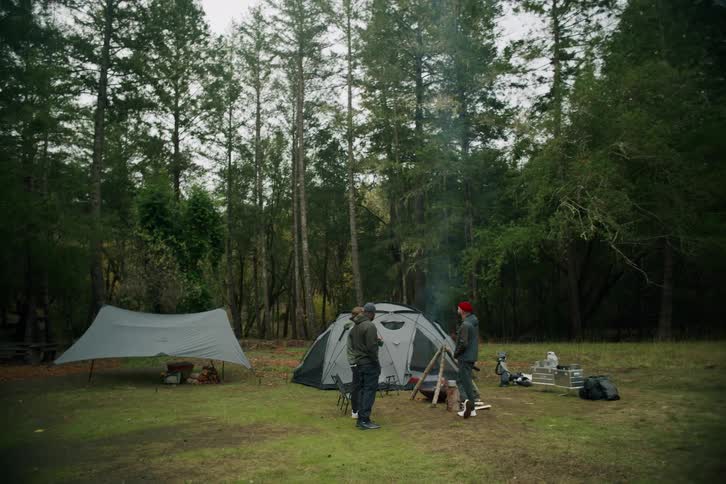 A group of friends enjoys a relaxing camping trip, standing around a smoldering campfire in a forest clearing