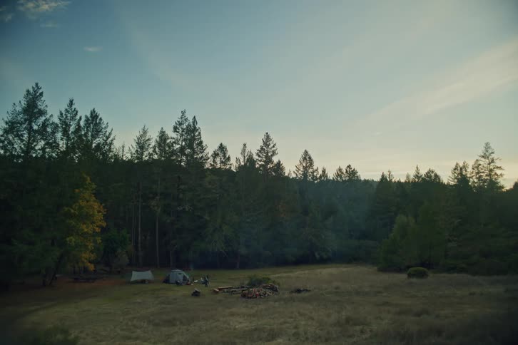 A wide shot of a tranquil campsite in a meadow bordered by a dense evergreen forest at dusk