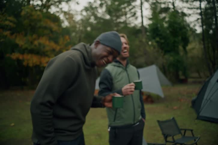 Close up on a steaming mug of coffee pulling out to reveal two diverse friends laughing together at their woodland campsite