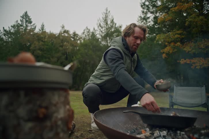 A man in a vest cooks a meal in a cast iron skillet over a fire pit while camping in the woods
