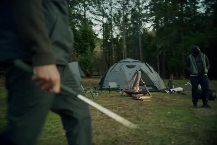 A man walks in slow motion through a campsite with a golf club and empties a bag of balls onto the grass to practice his swing