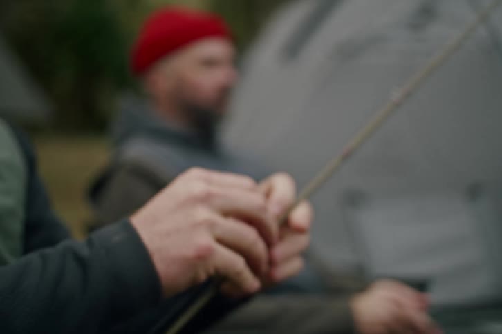 A fisherman carefully threads the line through his fishing pole with a tent visible in the background