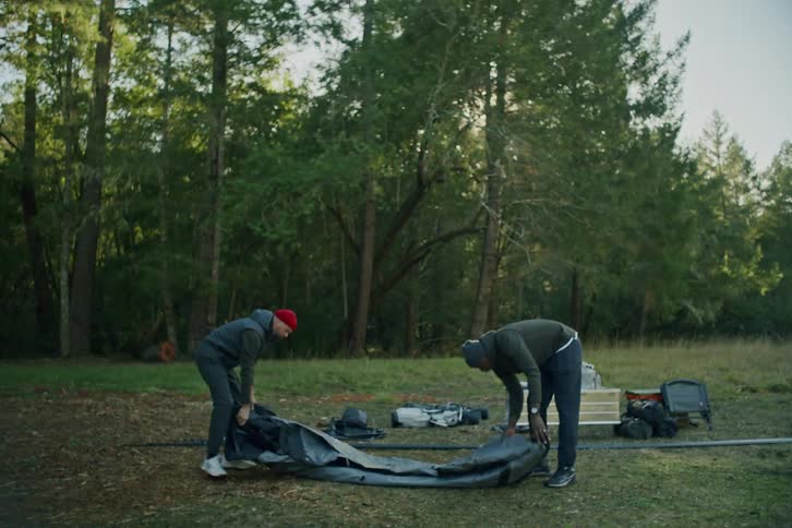 Two male friends work together in slow motion to set up a large tent in a forest clearing for their camping trip