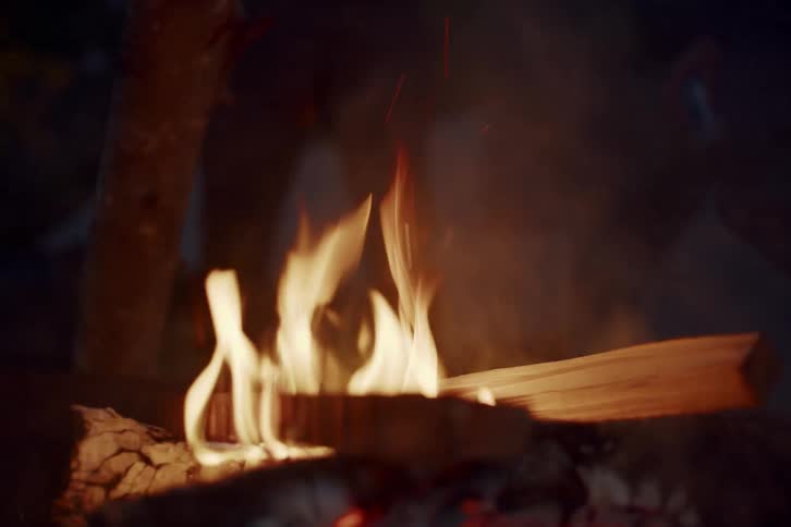 A person carefully places a piece of wood on a roaring campfire at night in slow motion, sending sparks flying