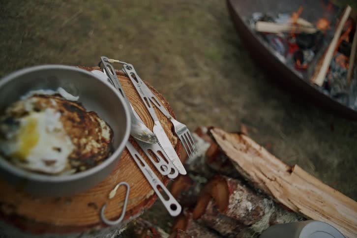 A rustic campsite meal with fried eggs in a bowl and cutlery rests on a tree stump next to a warm, crackling fire pit