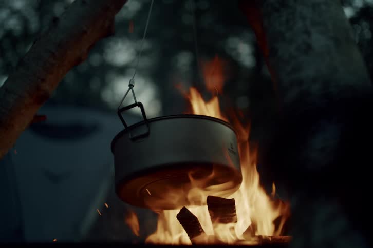 A metal cooking pot hangs over a crackling campfire at a campsite in the evening, with sparks flying in slow motion