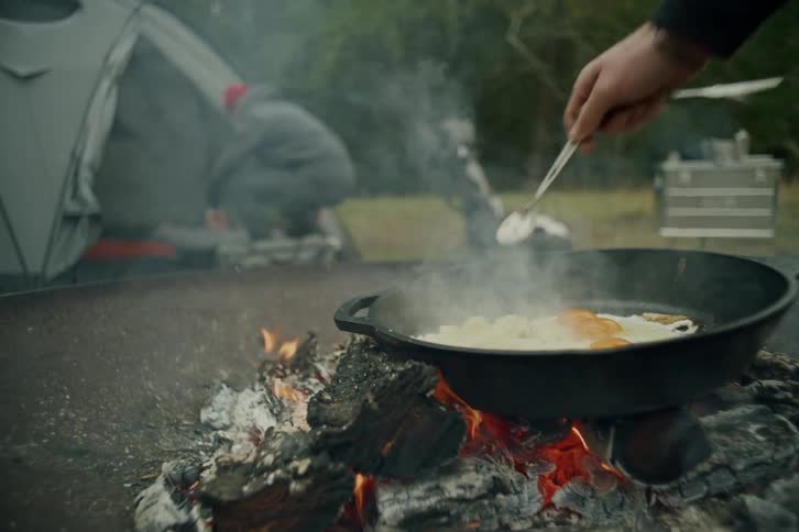 A person cooks a hearty breakfast of eggs and potatoes in a cast iron skillet over a campfire at their campsite
