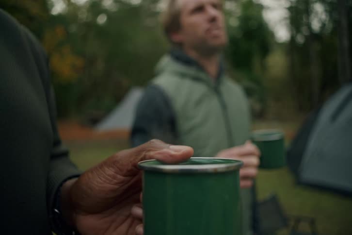 Two male friends enjoy a hot drink in green enamel mugs while camping together in the woods