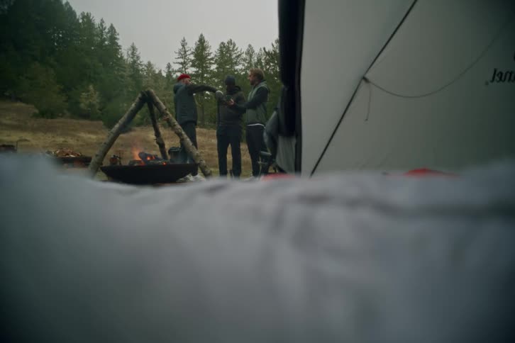 Three friends gather around a campfire at their campsite, talking and drinking from mugs on an overcast day