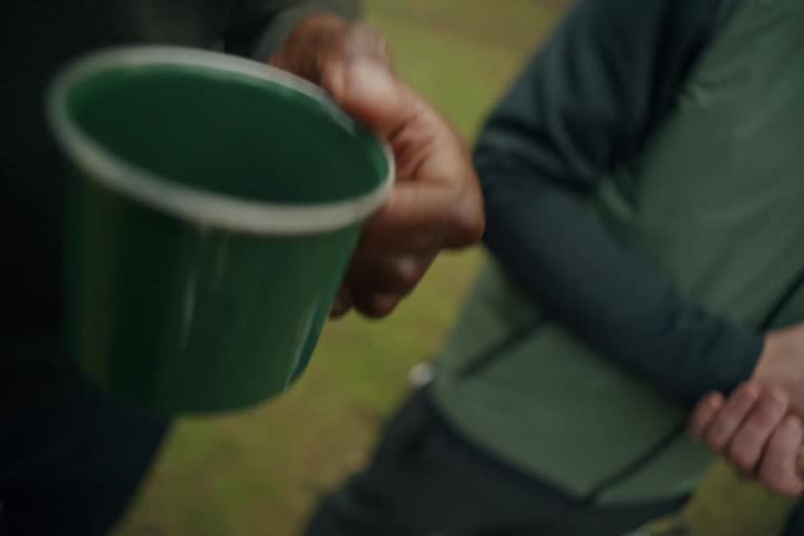 A close-up slow motion shot of a person's hand bringing a green enamel camping mug of coffee up for a drink while relaxing outdoors