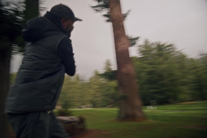 Slow motion shot follows a man as he tees off with a powerful swing on a golf course surrounded by towering evergreen trees