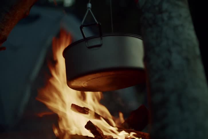 A lightweight camping pot hangs over a blazing bonfire at a campsite as the camera slowly tilts down to the flames