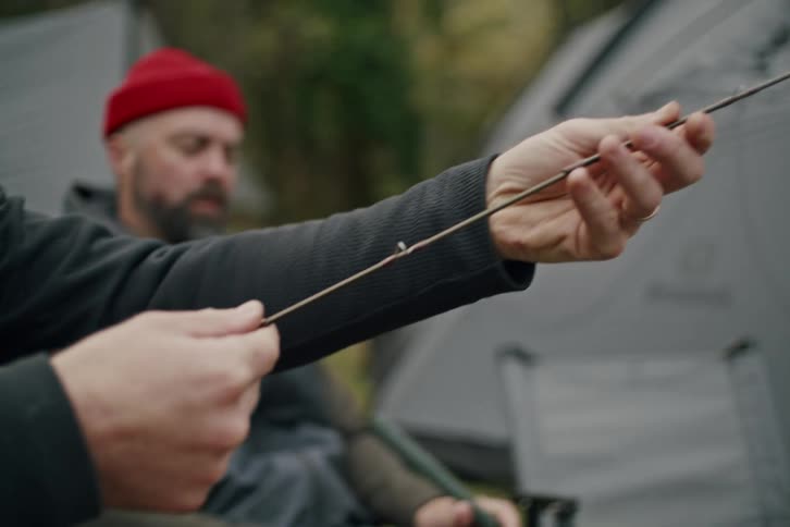 Close up of a man's hands assembling a multi-piece fishing rod at a campsite with his friend in the background