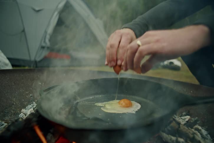 Slow-motion shot of a man cracking an egg into a smoking cast-iron skillet over a campfire at a campsite