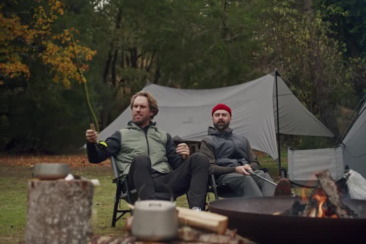 Two male friends relax at their forest campsite, one tending to his fly fishing gear while the other roasts a marshmallow over the fire