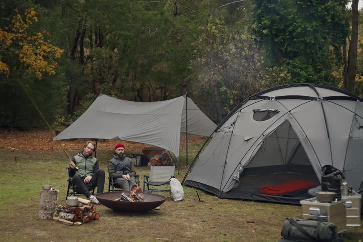 Two male friends sit around a fire pit talking and relaxing at their campsite in the woods