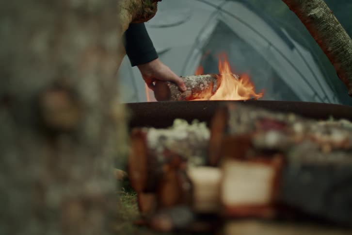 A camper's hand grabs firewood and places it on a crackling fire next to their tent in the woods