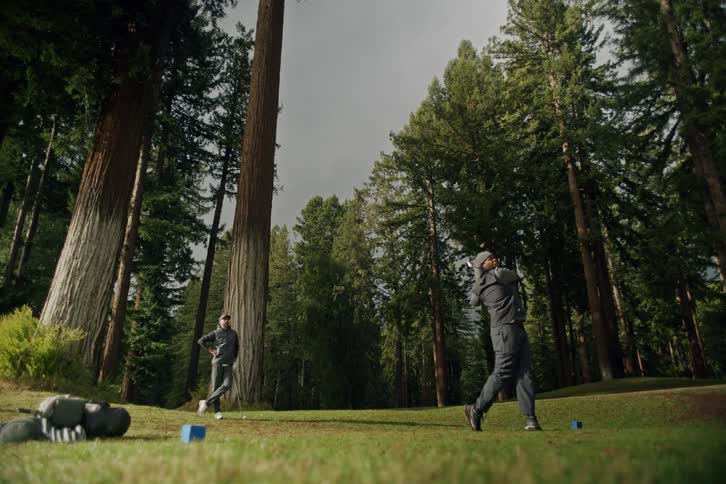 A man takes a powerful golf swing in slow motion on a magnificent course surrounded by towering redwood trees as his friend watches