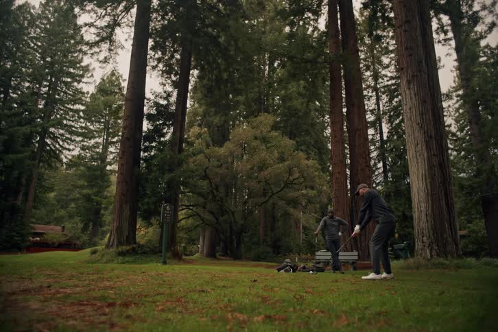 A man takes a slow-motion golf swing and flips his club on a course surrounded by giant redwood trees