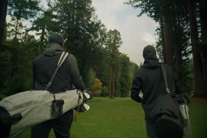 A slow-motion tracking shot from behind two men carrying their golf bags as they walk down a scenic fairway surrounded by towering redwood trees