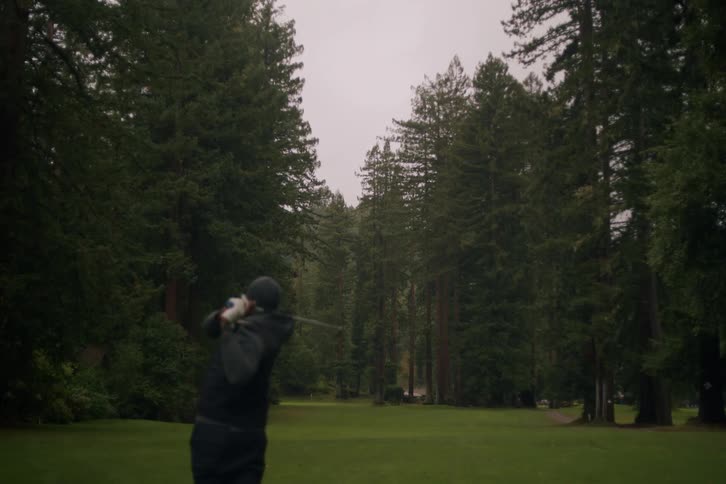 A man in a beanie and vest takes a powerful swing with his golf club in slow motion on a green course surrounded by tall redwood trees