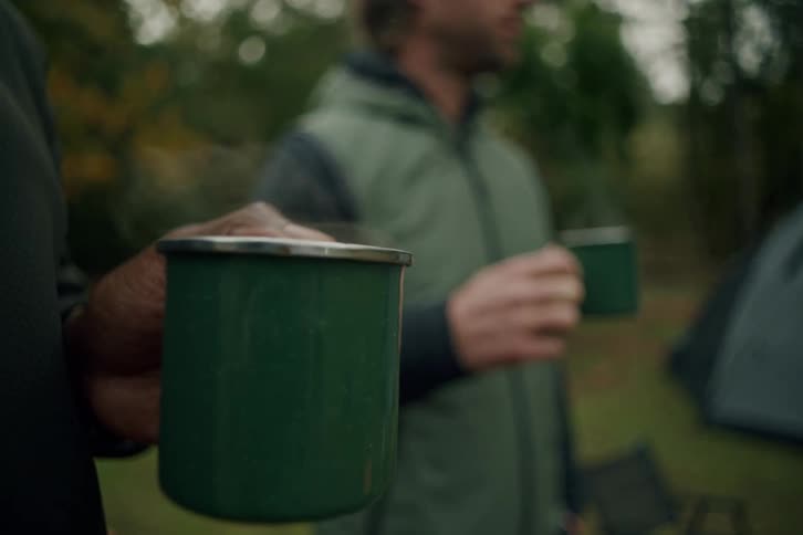 Slow motion close-up of two men holding steaming green enamel mugs while camping in the woods