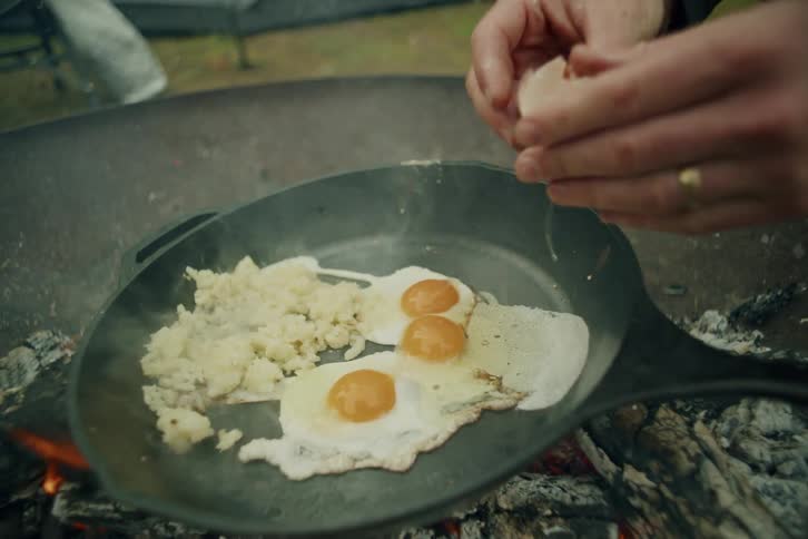 Slow motion shot of a person cracking a fresh egg into a sizzling cast iron skillet of potatoes and eggs over a campfire
