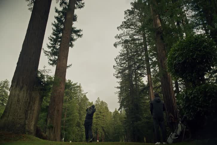 A golfer tees off in slow motion on a beautiful course surrounded by towering redwood trees while his friend watches