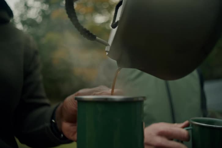 Slow motion shot of a man pouring hot coffee into mugs for his friends at their campsite in the woods