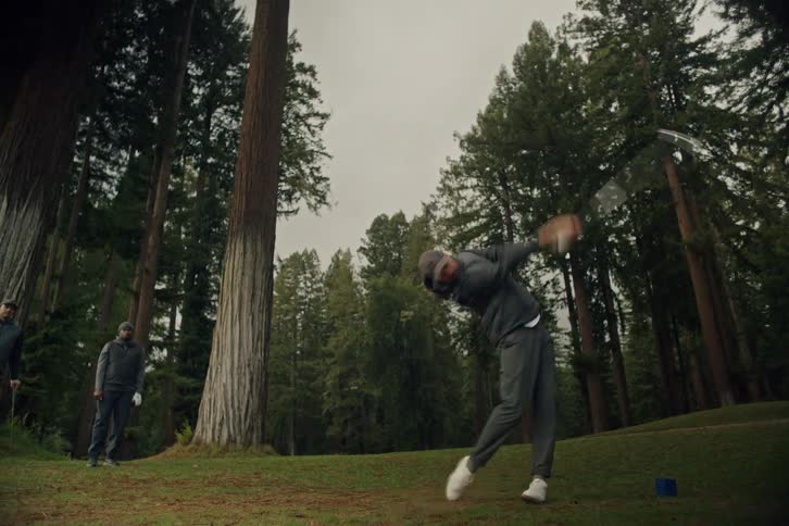 A male golfer takes a powerful slow-motion swing on a scenic golf course surrounded by giant redwood trees