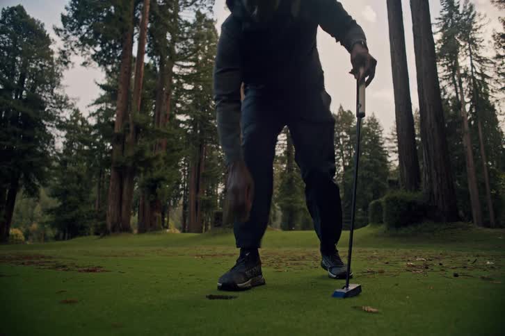 A man sinks a putt on a scenic golf course surrounded by towering redwood trees