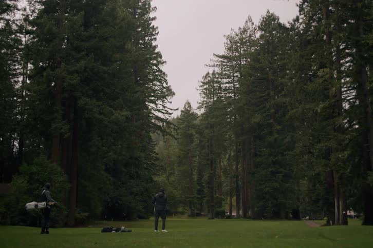 A man practices his golf swing on a beautiful fairway surrounded by towering redwood trees as his caddie looks on