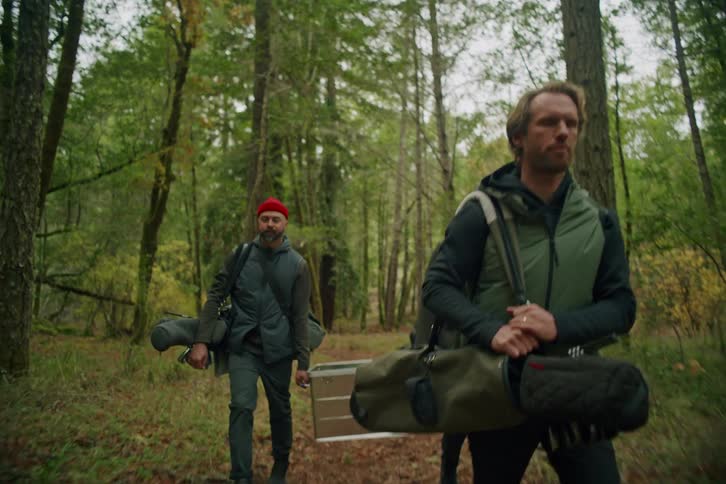 Three diverse male friends hike through a dense forest carrying golf bags and a gear case on an adventure